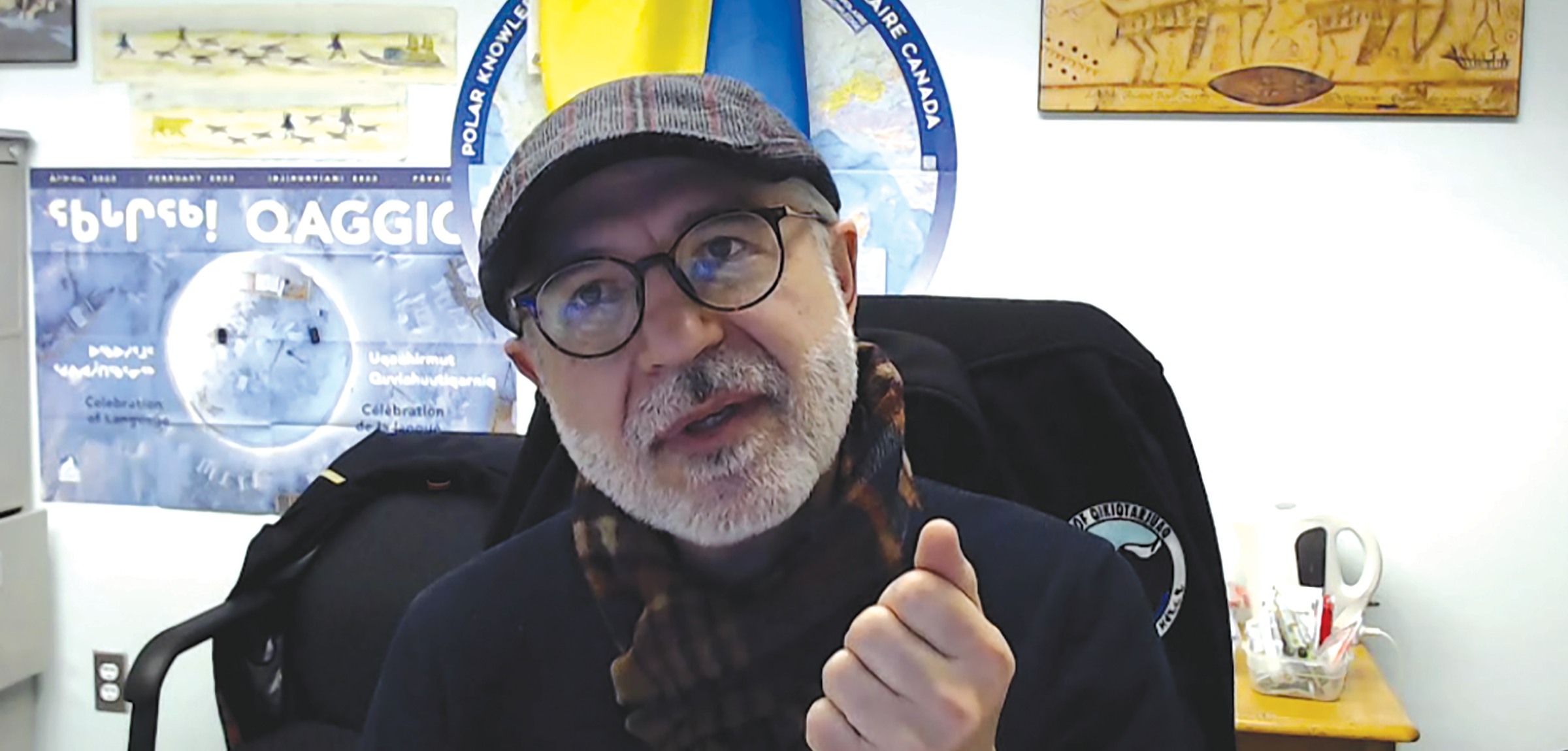 A older, bearded man wearing glasses, a cap, and a scarf speaks from his desk in front of a Ukrainian flag