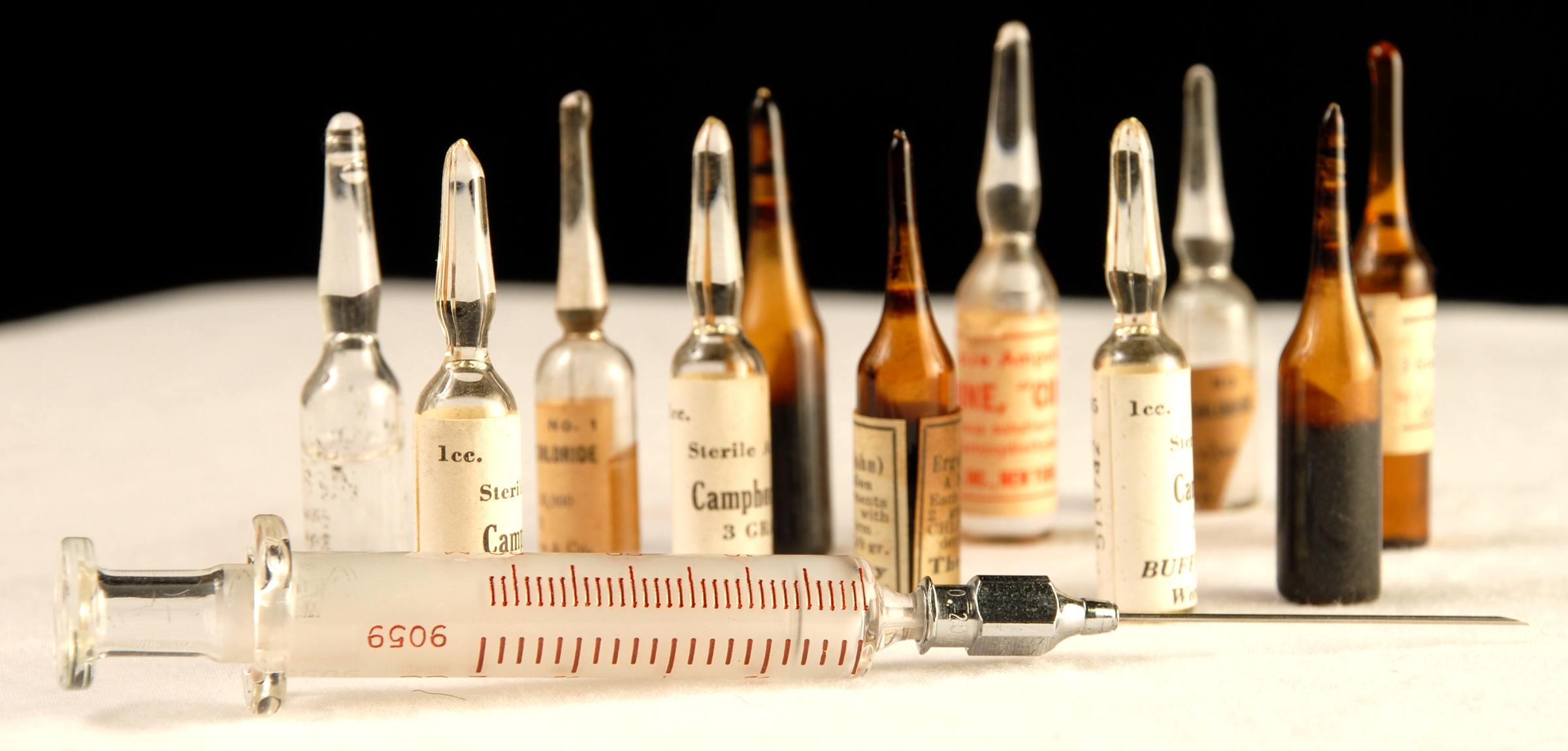 An empty syringe lies sideways in front of a variety of glass vials