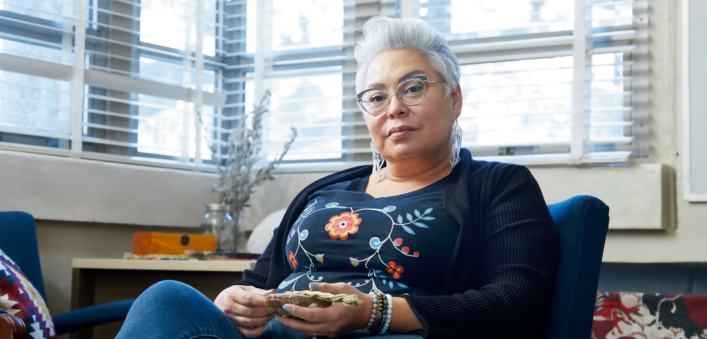 Janis Wasel Bear, an older Indigenous woman with short grey hair and glasses, sits in her office and looks directly into the camera