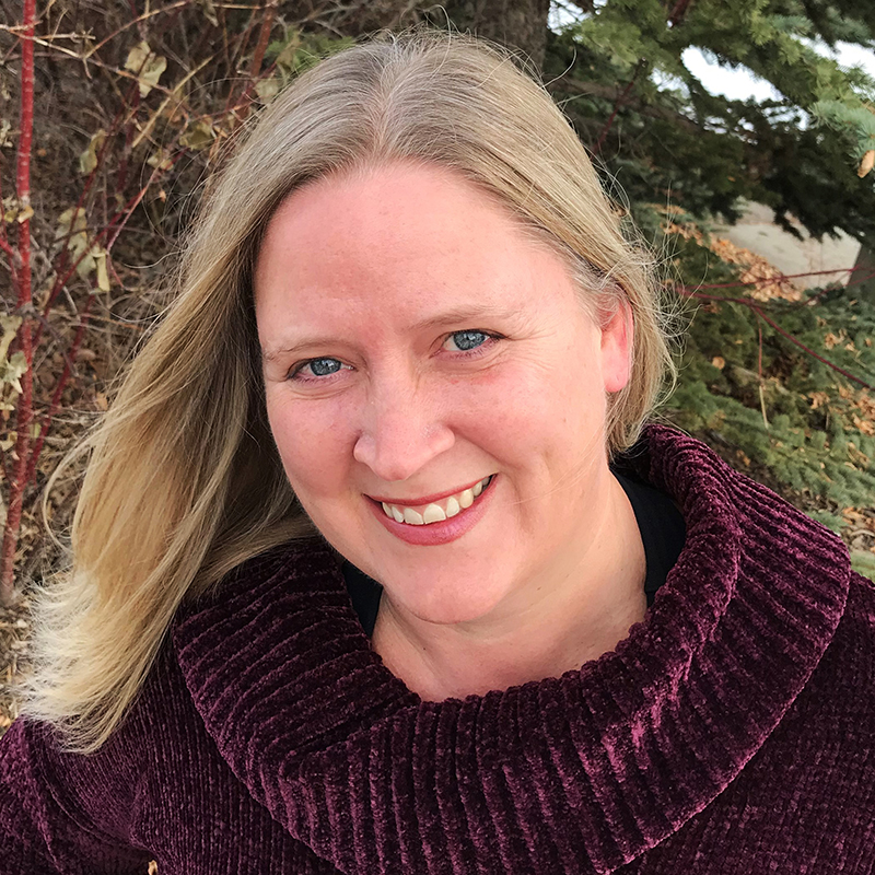 White women with blonde hair standing in front of a spruce tree wearing an eggplant coloured turtleneck sweater