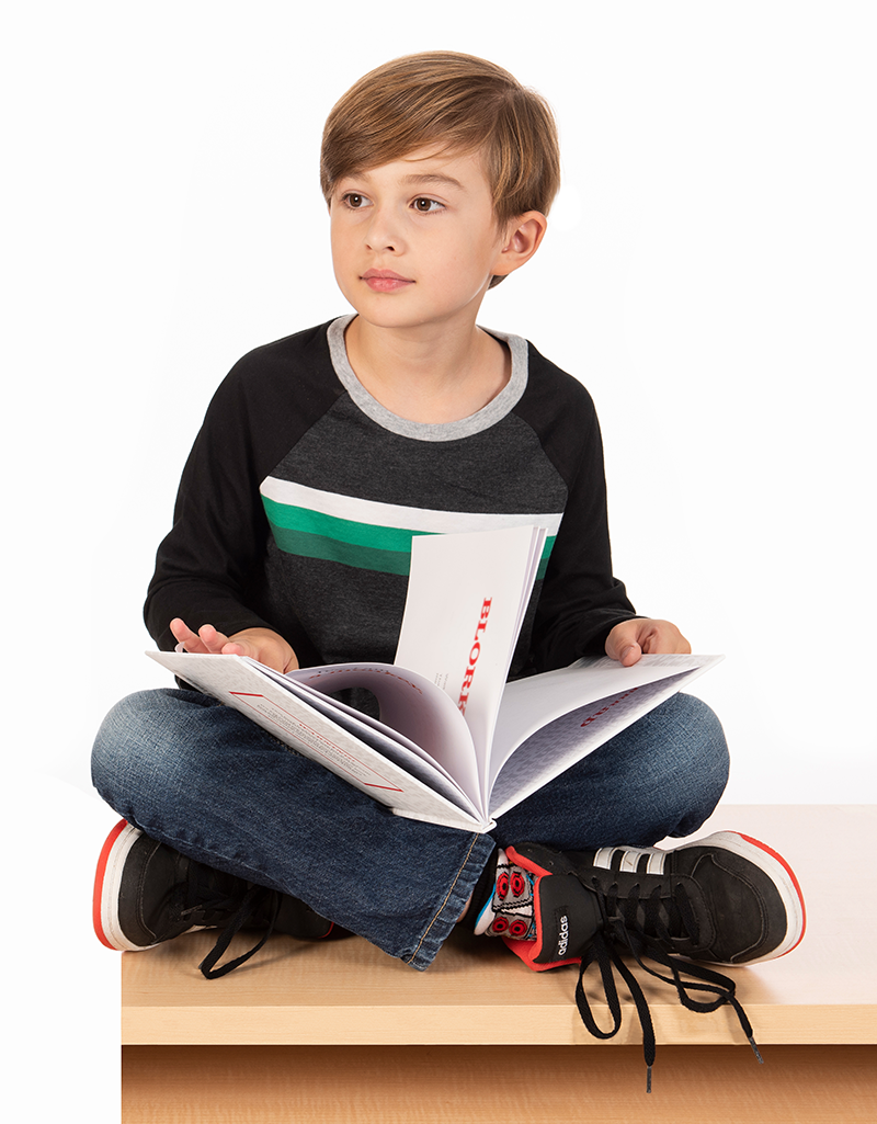 White boy with baseball shirt sits cross-legged on a table with a book in his lap.