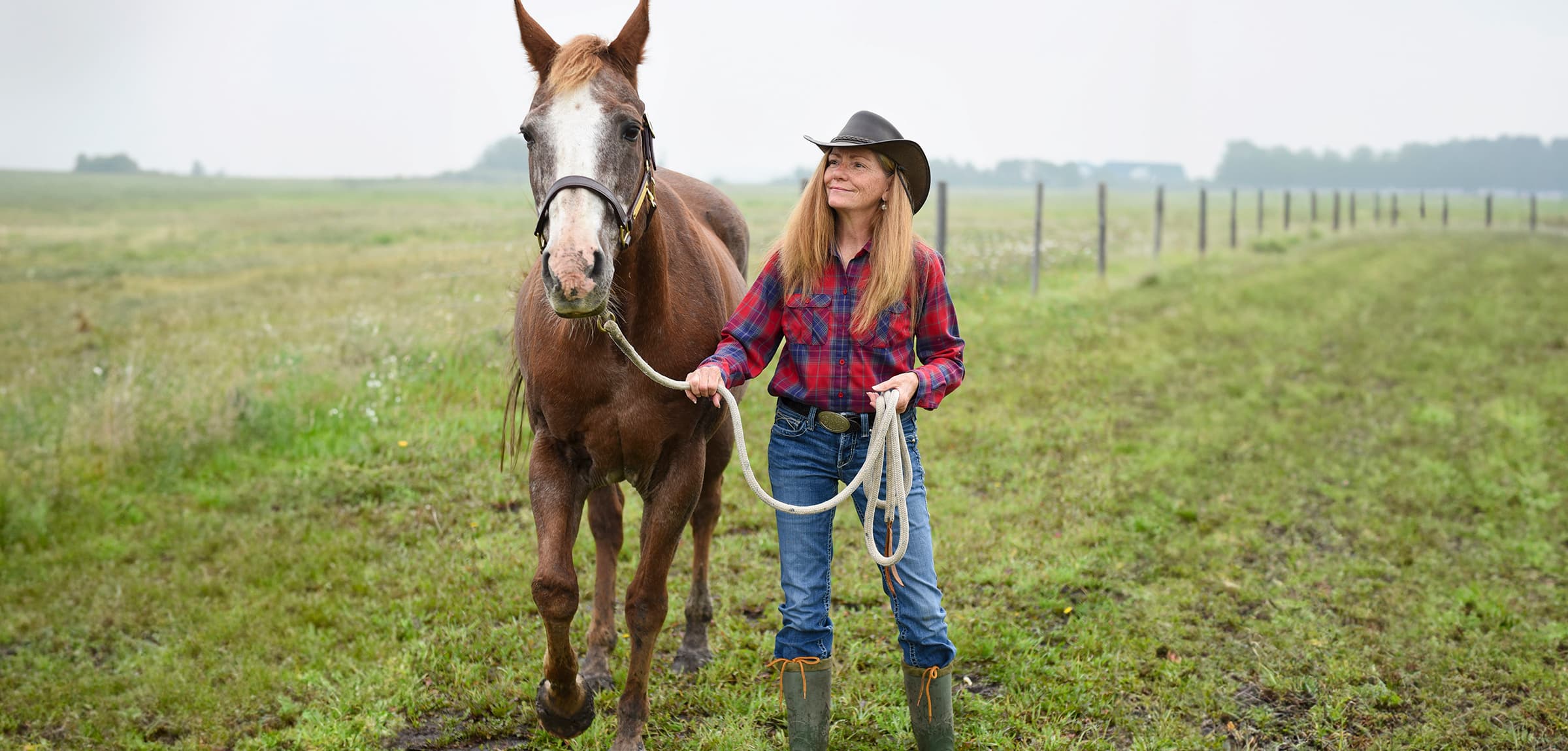 Women in a cowboy had standing in a field leading a chestnut coloured horse