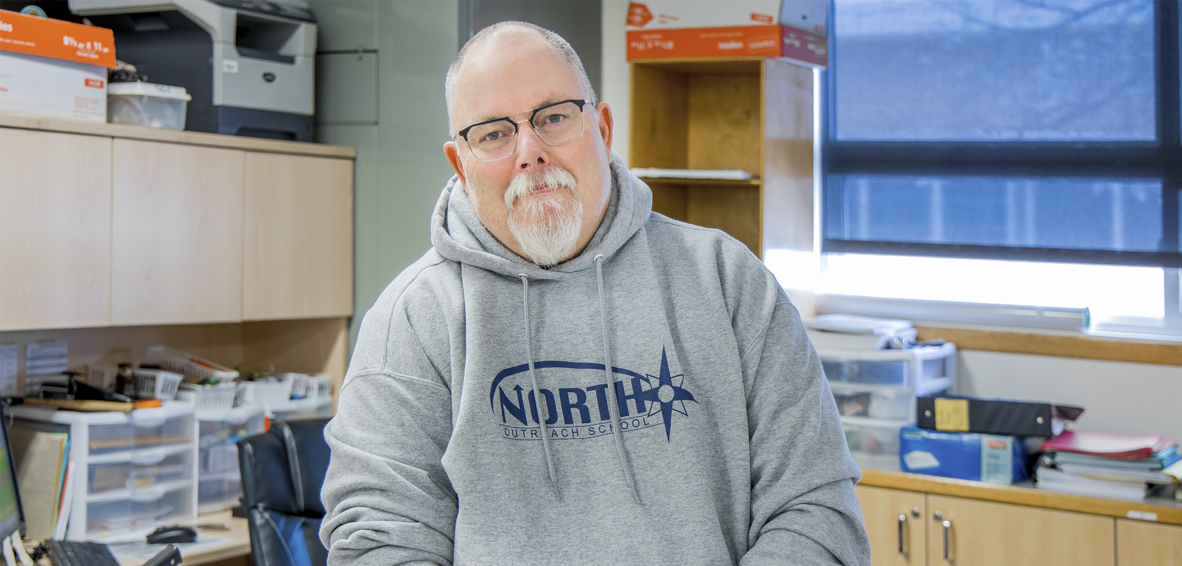 A man with short gray hair and glasses wearing a gray hoodie and sitting on a desk