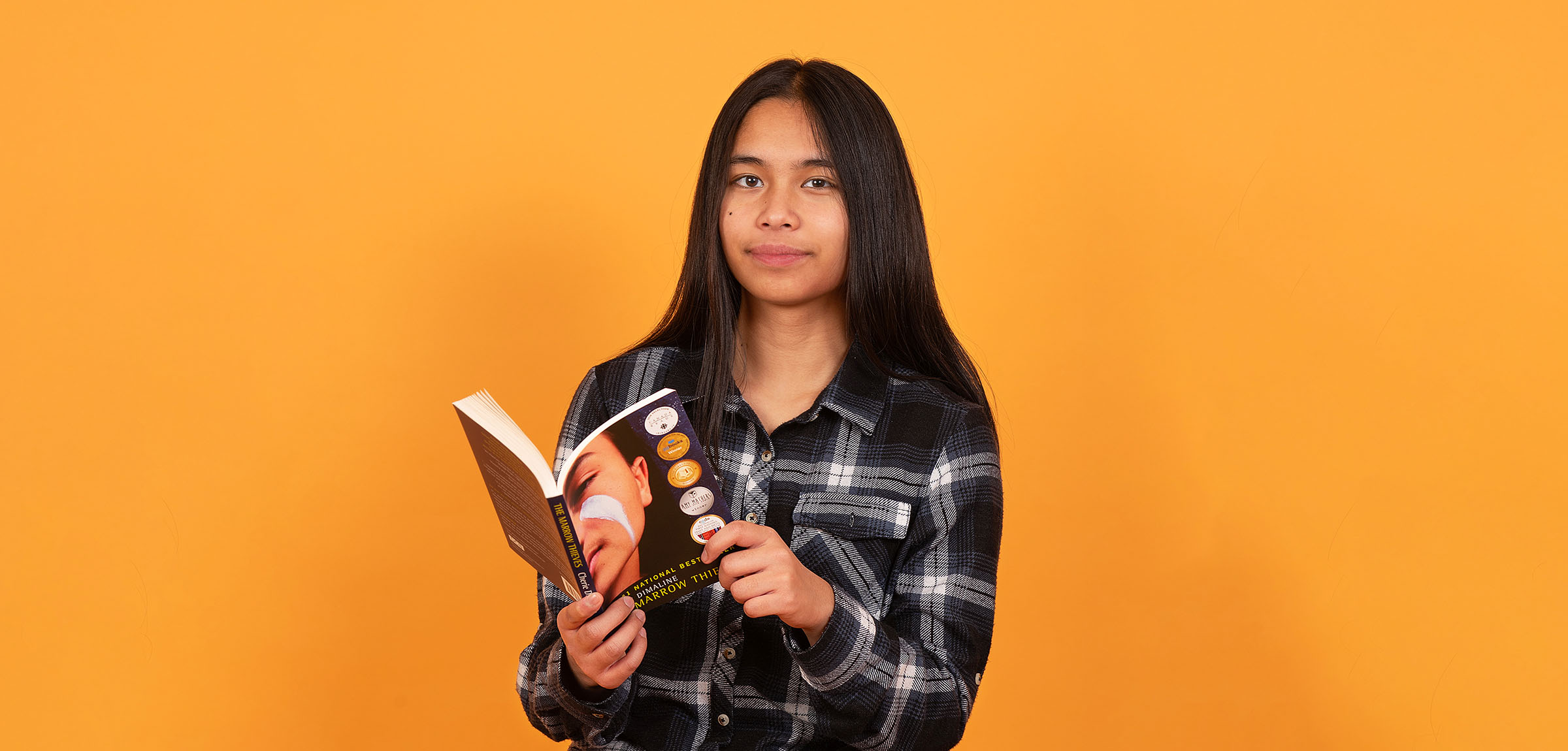 Boy with long dark hair wearing a black plaid shirt reading a book infront of an orange wall