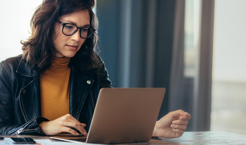 Woman in leather jacket and yellow shirt looking at laptop next to a window