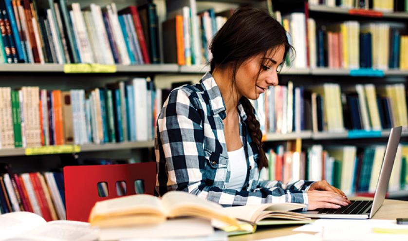 Young women of colour sitting at a table in a library writing with a pencil in front of an open laptop. 