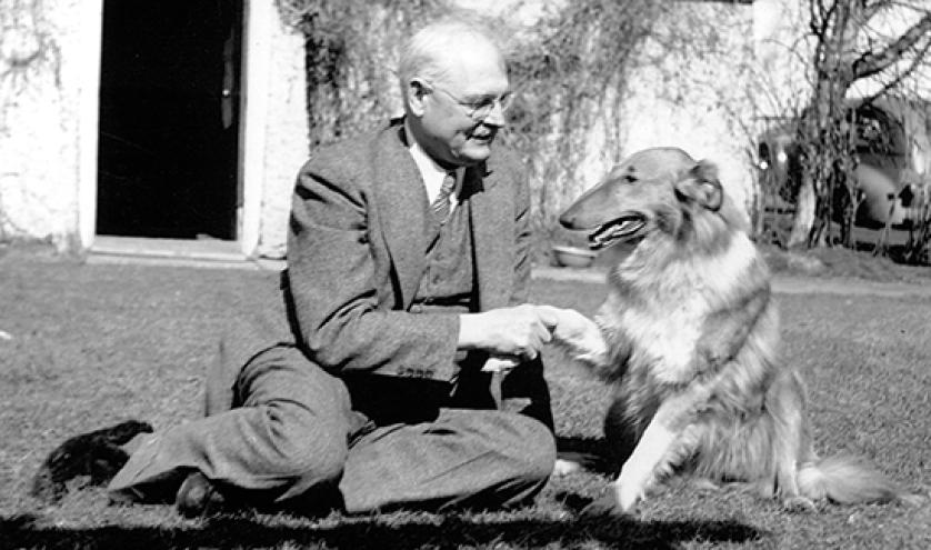 black and white photo of a caucasian man seated on the ground in a suite, shaking the paw of a rough collie dog.