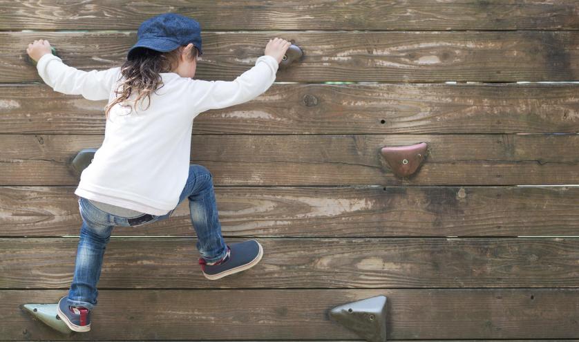 Young child climbing a wall with grips wearing blue jeans, a hat and white shirt