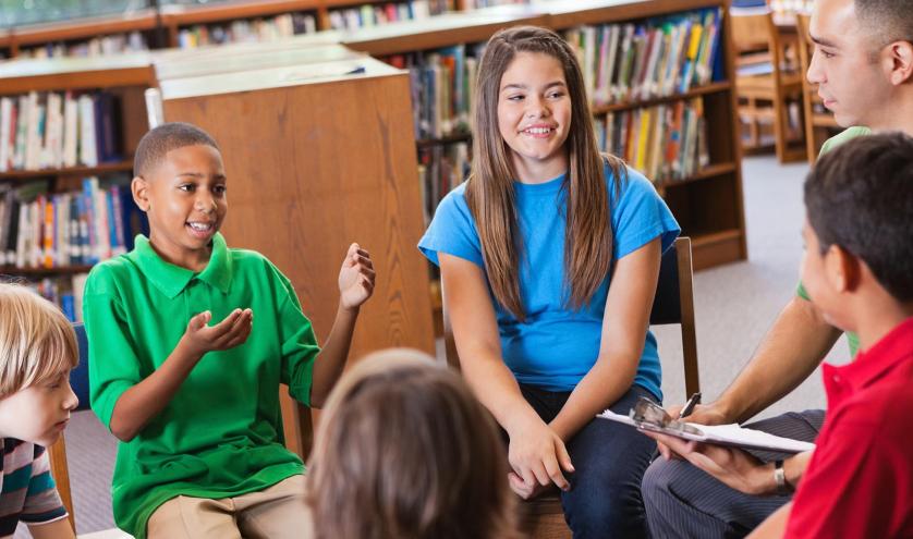 Students and their teacher seated in chairs listening to a young boy speak