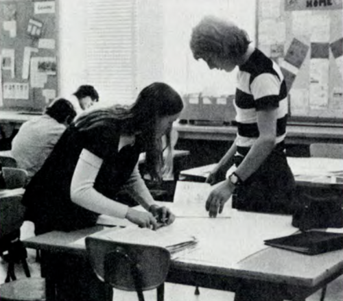 A black and white image of a teacher dressed in a striped shortsleeve shirt standing at a student's desk.