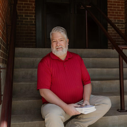 Man with a grey beard and hair sits on stairs in a red shirt