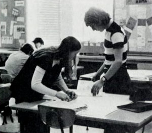 A black and white image of a teacher dressed in a striped shortsleeve shirt  standing at a student's desk.