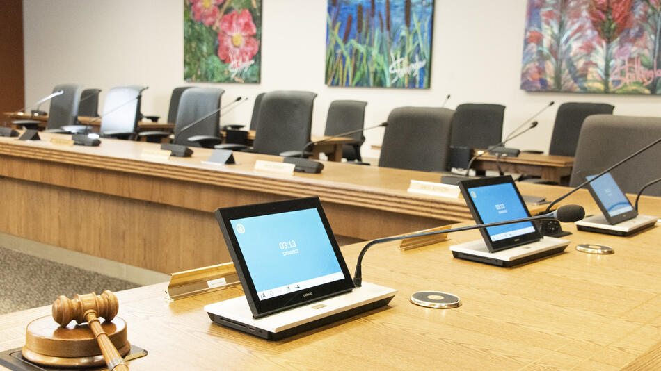 Provincial Executive Council chamber a large round wooden desk lined with microphones and chairs facing the center and paintings of nature on the wall.