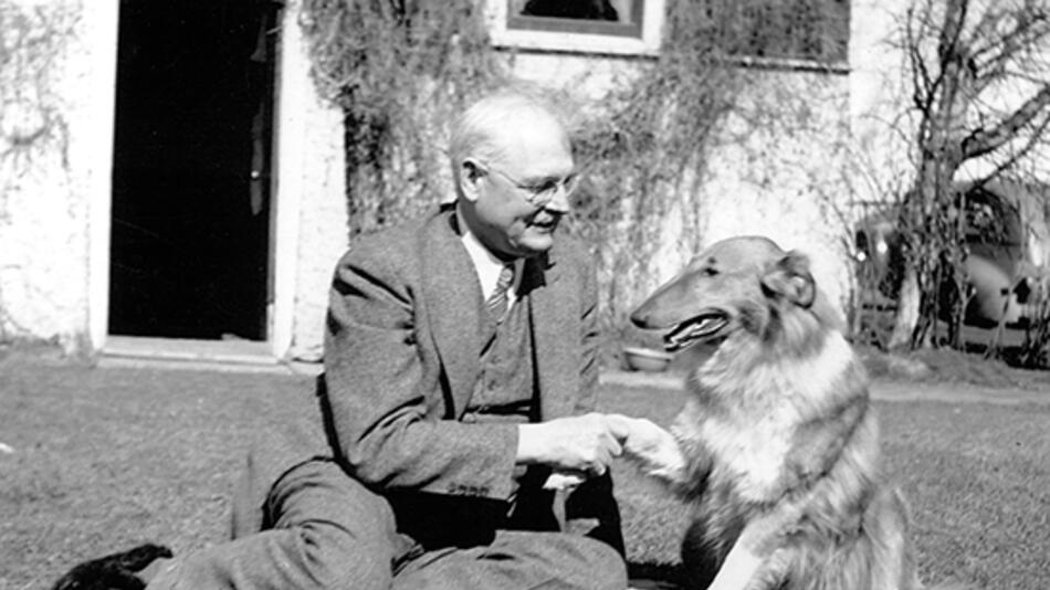 black and white photo of a caucasian man seated on the ground in a suite, shaking the paw of a rough collie dog.