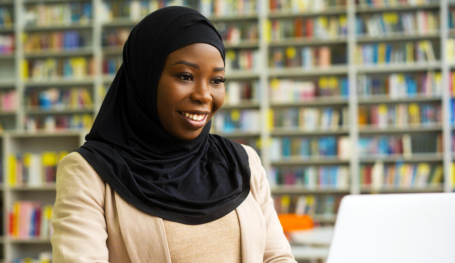 A woman in a hijab using a laptop in a library. 