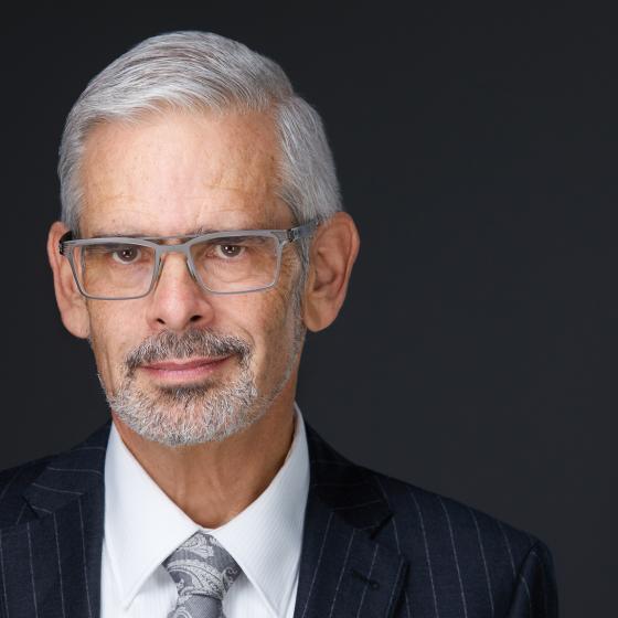 Dennis Theobald with silver white goatee and hair wearing a dark suit and tie in front of a black background.