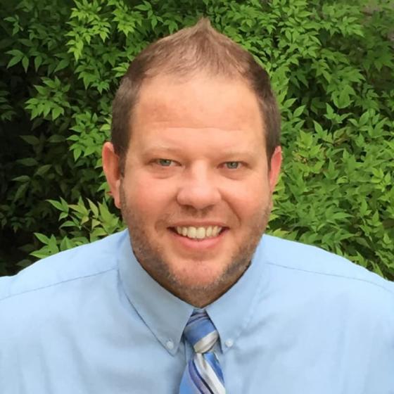 Portrait of middle age Caucasian man wearing a blue shirt and tie standing in front of a green bush