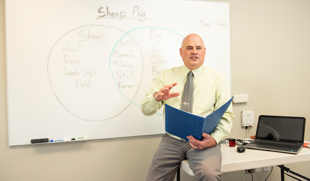 White bald man sitting at the edge of a desk in a pale yellow shirt, holding a blue binder, speaking in front of a white board with a Venn diagram describing a sheep and a pig.