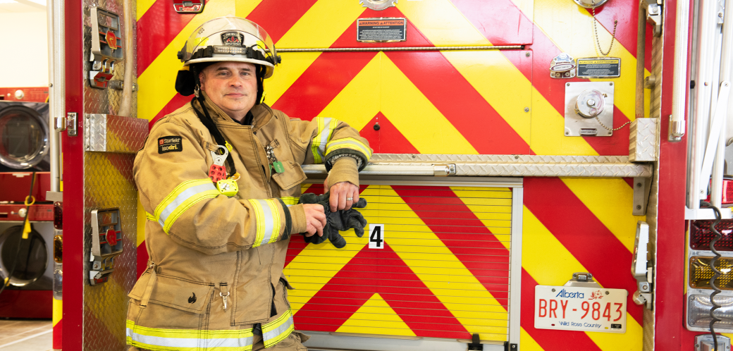 Firefighter standing in full uniform leaning on the back of a fire truck with a high contrast yellow and red chevron pattern.