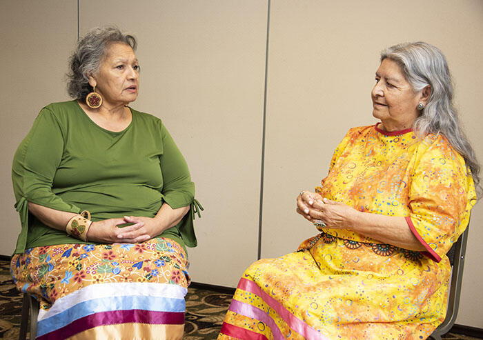 Two elders wearing ribbon skirts having a seated conversation.