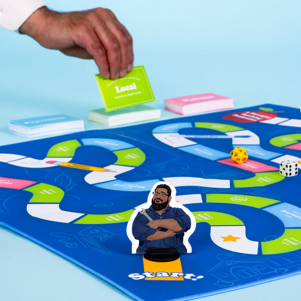 Get started A photograph of a bright blue boardgame on a light blue background. In the background, a man's hand pick up playing cards that say "Local volunteer opportunity". In the foreground, a player token is at the start of the boardgame. The player is an illustration of D'Souza in his mechanic outfit, crossing his arms confidently and holding a wrench.