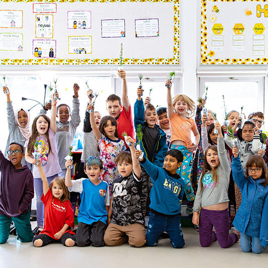 A classroom of children hold up tree seedlings