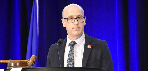 ATA president Jason Schilling stands on stage behind a podium in a suite and tie