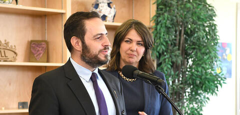 Man with a beard speaks at a microphone with a women standing behind him in a navy suit