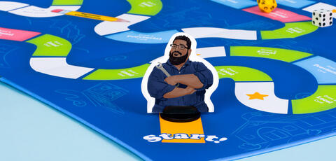 A photograph of a bright blue boardgame with a focus on a player token at the start of the game. It is an illustration of D'Souza in his navy blue mechanic suit, holding a wrench and smiling.