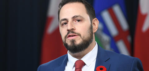 Education Minister infront of flags wearing a poppy in a blue suit