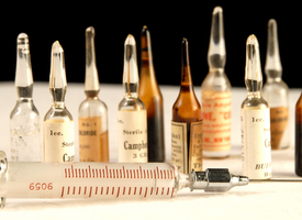 An empty syringe lies sideways in front of a variety of glass vials