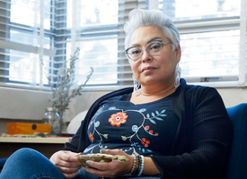 Janis Wasel Bear, an older Indigenous woman with short grey hair and glasses, sits in her office and looks directly into the camera