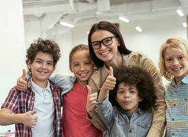 Teacher wearing glasses standing with 4 children all giving a thumbs up