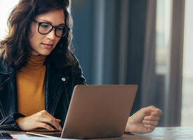 Woman in leather jacket and yellow shirt looking at laptop next to a window