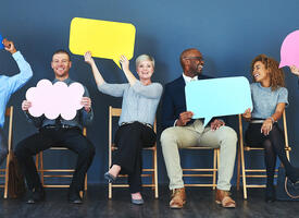 Group of adults sitting on chairs holding blank speech bubbles made of coloured paper
