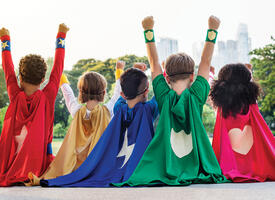 5 children faced away from the camera wearing different coloured superhero capes with hearts, lightning bolts and shields on the back.