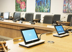 Provincial Executive Council chamber a large round wooden desk lined with microphones and chairs facing the center and paintings of nature on the wall.