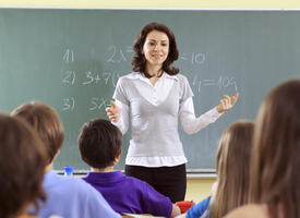 White female with brown hair standing in front of a chalkboard teaching students in the foreground 