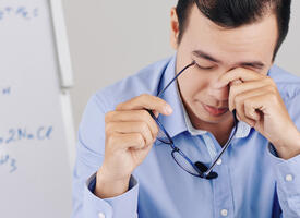 Middle age man rubbing holding his glasses and rubbing his eyes infront of a white board with equations