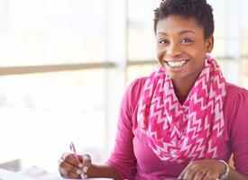 Women with short black hair wearing a fuscia scarf and sweater smiling while working at a desk.