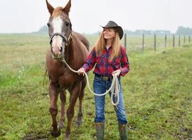 Women in a cowboy had standing in a field leading a chestnut coloured horse