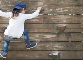 Young child climbing a wall with grips wearing blue jeans, a hat and white shirt