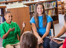 Students and their teacher seated in chairs listening to a young boy speak