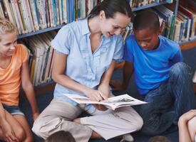 Female teacher seated on library floor with a group of students.