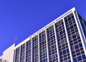 A blue sky background free of clouds. In the foreground is a white building with columns of blue windows. The canadian flag flies from the top of the building. The ATA logo and the text "Alberta Teachers' Association" are also on the front facade.