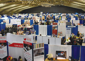 A photo of the inside of a large convention centre. The top of the photo is yellow with beams of the roof and lights. On the floor are people, many displays and blue dividers for the booths.