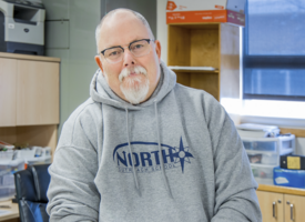 A man with short gray hair and glasses wearing a gray hoodie and sitting on a desk