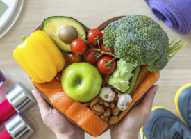 The background is a light wood table or floor. There is a scale, a yoga mat, weights, and running shoes in each corner. In the foreground two hands hold fruits, vegetables, nuts and salmon. These items of food are arranged in a heart shape. 