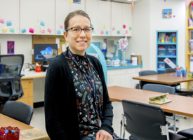 A classroom with white walls and brown tables for desks. There is bunting around the classroom and colourful sitckers on the white cupboards. In the foreground a brunette sits wearing a black dress speckled with colour and a black cardigan above it. 