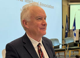 Grey haired man stands at a podium in front of an audience.